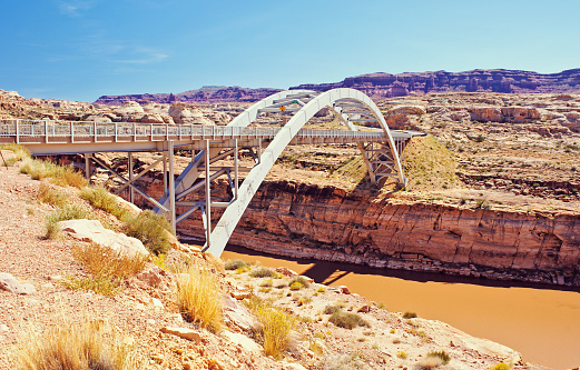The Hite Crossing Bridge is an arch bridge following Route 95 across the muddy Colorado River in the Utah desert. Different layers of sedimentary rock are visible in the cliffs where the river has carved a canyon below. Desert mesas can be seen off in the distance.