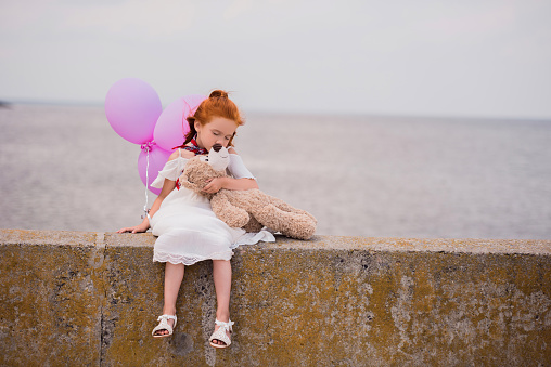 adorable little girl with teddy bear and balloons sitting on quay