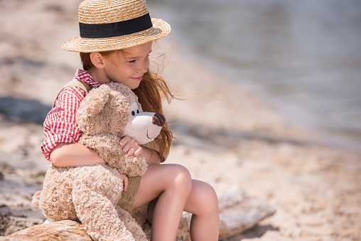 pensive little girl hugging teddy bear and looking away while sitting on seashore
