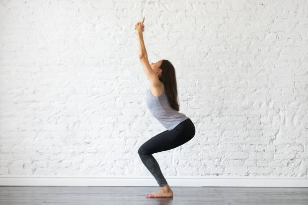Young attractive woman in Chair pose, studio background Young attractive woman practicing yoga, standing in Chair exercise, doing Utkatasana pose, working out, wearing sportswear, gray tank top, black pants, indoor full length, studio background cat pose yoga stock pictures, royalty-free photos & images