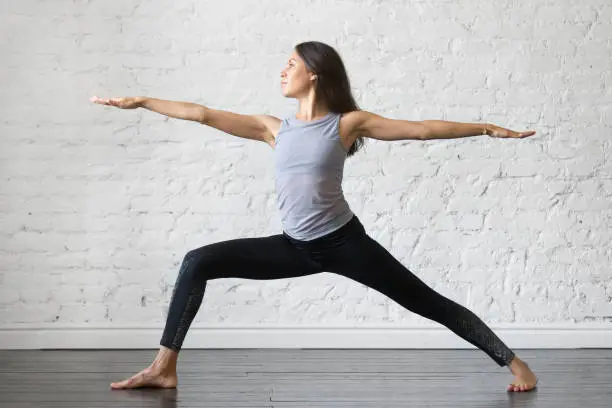 Young attractive woman practicing yoga, standing in Warrior one exercise, Virabhadrasana II pose, working out, wearing sportswear, gray tank top, black pants, indoor full length, studio background