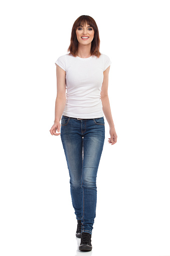 Happy young woman in jeans and white t-shirt is walking towards camera and talking. Front view. Full length studio shot isolated on white.