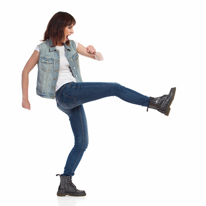 Young woman in jeans vest and black boots is shouting and kicking. Side view. Full length studio shot isolated on white.