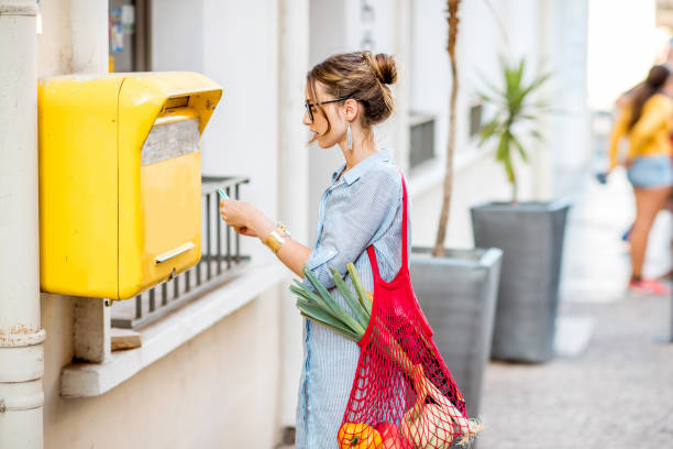 Woman using old mailbox outdoors Young woman putting letter to the old yellow mailbox standing with mesh bag full of food outdoors on the street post office stock pictures, royalty-free photos & images