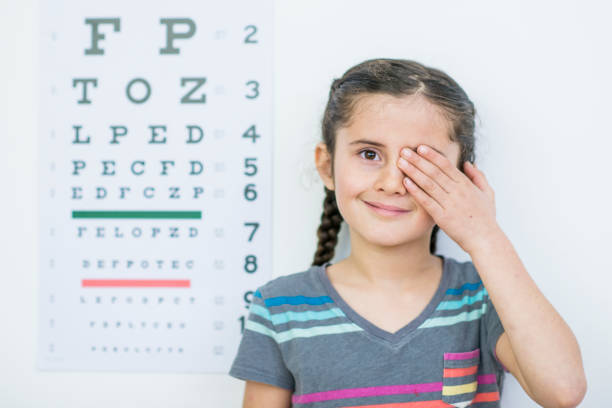 Eye Closed An Ethnic girl is indoors in an optometrist's office. She is wearing casual clothing. She is standing beside an eye chart and closing one eye for a test. eye test equipment stock pictures, royalty-free photos & images
