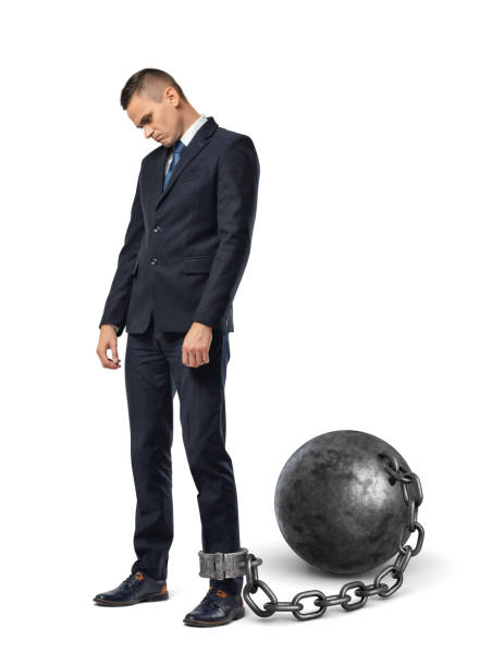 a sad businessman looks down while shackled to a large iron ball with a chain to his ankle - isolated businessman sadness business person imagens e fotografias de stock