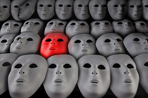 Glowing red face mask among many white masks on black background. Alone in the crowd concept