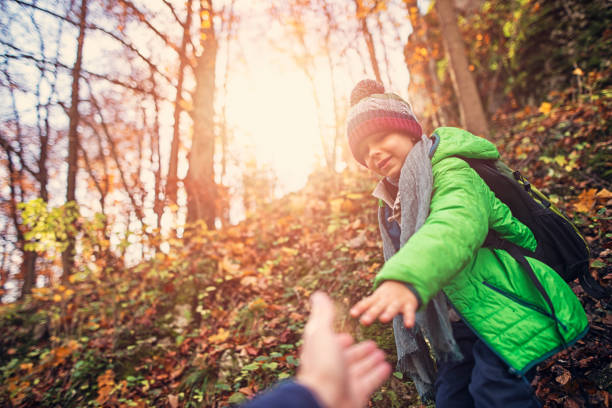 Father and son are hiking in autumn forest. Fother is helping the son to walk down very steep slope. Shot from father's perspective as he reaches for the son's hand.