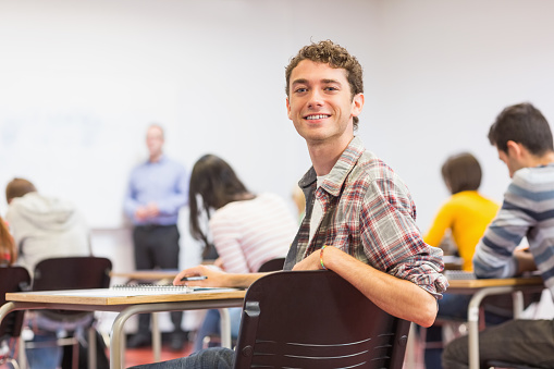 Portrait of a smiling young male student with blurred teachers and others in the classroom