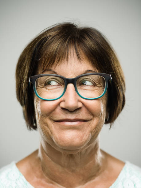 Real senior woman portrait looking away Close up portrait of hispanic mature woman with naughty expression against gray background. Vertical shot of real senior woman with happy smile and looking to the side in studio. Short brown hair and colorful modern glasses. Photography from a DSLR camera. Sharp focus on eyes. looking around stock pictures, royalty-free photos & images