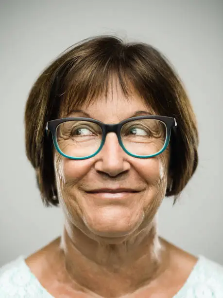 Close up portrait of hispanic mature woman with naughty expression against gray background. Vertical shot of real senior woman with happy smile and looking to the side in studio. Short brown hair and colorful modern glasses. Photography from a DSLR camera. Sharp focus on eyes.