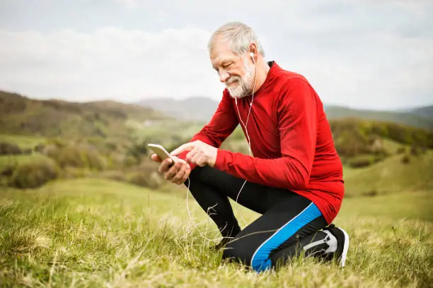 Active senior runner outside on green hills with smart phone and earphones, resting, listening music.