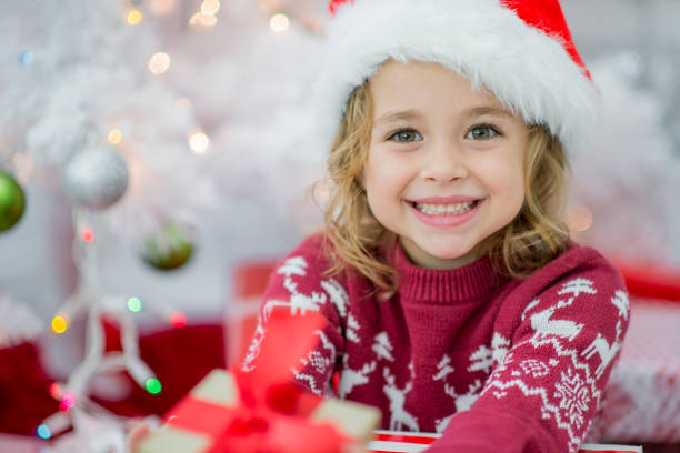 Excited about Christmas A young girl of elementary age is sitting under a white Christmas tree with lights and presents behind her. She is wearing a Santa hat and smiling up at the camera. surgical light stock pictures, royalty-free photos & images