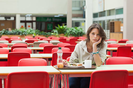 Portrait of a alone and sad female student sitting in the cafeteria with food tray