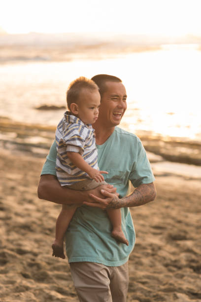 Eurasian father holds adorable son as they take an evening walk along a sandy Hawaii beach
