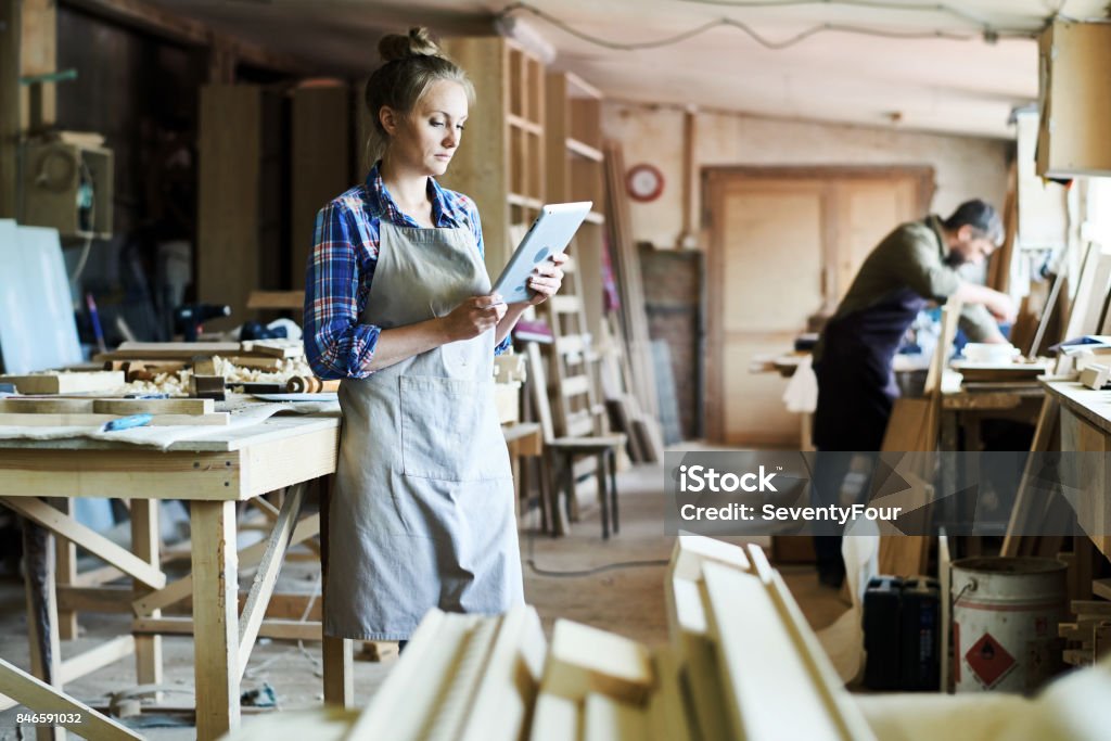 Attractive Craftswoman Using Digital Tablet Full length portrait of concentrated young craftswoman in apron standing at spacious workshop and using digital tablet Carpenter Stock Photo