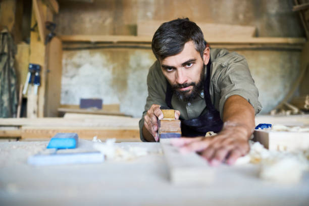 Handsome Craftsman Planing Plank Concentrated bearded craftsman wearing apron smoothing plank with jointer plane, shavings scattered on table carpenter portrait stock pictures, royalty-free photos & images