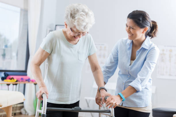 Healthcare professional helps senior woman walk with a walker Cheerful senior Caucasian woman uses a walker during physical therapy session. A mid adult Asian female physical therapist is helping her walk with the walker. infarction photos stock pictures, royalty-free photos & images