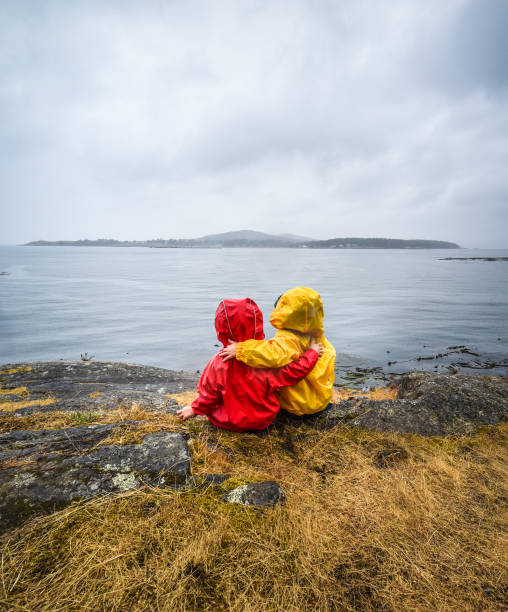 Two children wearing rain suits put their arms around each other while looking out at the ocean. Lopez Island, Washington State, USA.