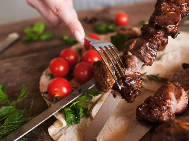 Piece of shish-kebab is taken off from skewer. Grilled meat, field mushrooms and cherry tomatoes on lavash, barbecue and natural food preparing in restaurant, close up picture
