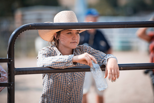 Young cowgirl is holding bottle of water and leaning on a fence in rodeo arena.