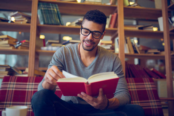 studente afroamericano sorridente che si gode la lettura di libri in biblioteca - reading book men sofa foto e immagini stock