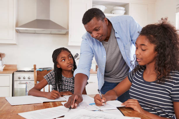 Father Helping Two Daughters Sitting At Table Doing Homework Father Helping Two Daughters Sitting At Table Doing Homework single father stock pictures, royalty-free photos & images