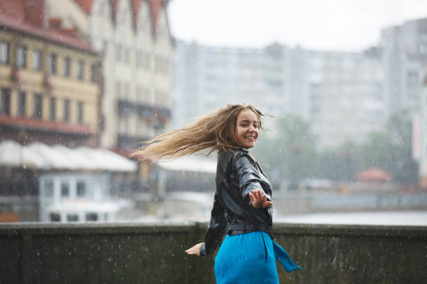 Waist up shot of a young smiling woman with black hair and in a raincoat looking up to the sky with her arms spread