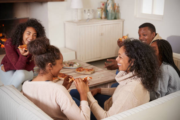familia sentada en el sofá en el salón junto a la chimenea, comiendo pizza - warming up fotos fotografías e imágenes de stock