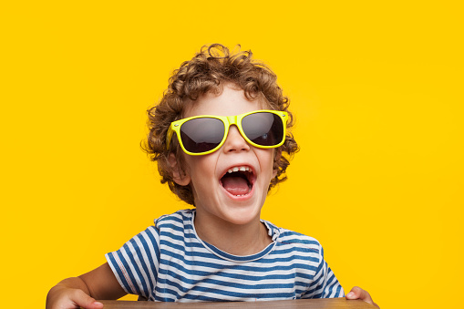 Portrait of cheerful little boy wearing adult sunglasses and looking away on orange background.