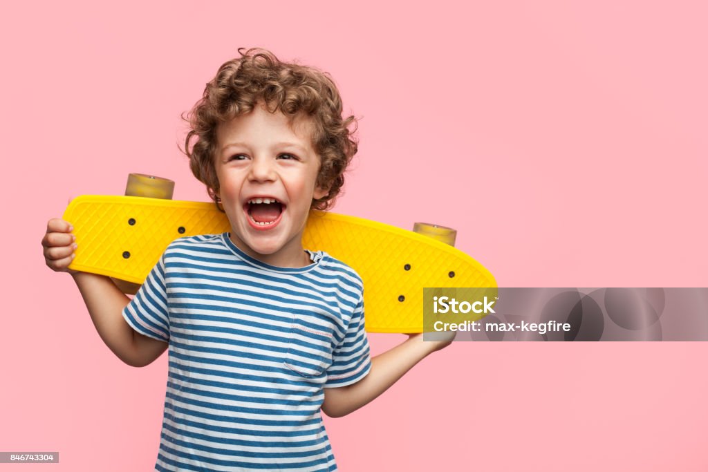 Cheerful boy with yellow longboard Charming curly boy holding yellow longboard and looking away on pink background. Child Stock Photo