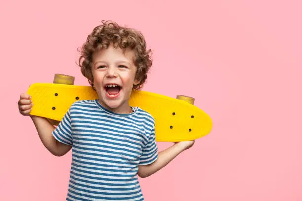 Charming curly boy holding yellow longboard and looking away on pink background.