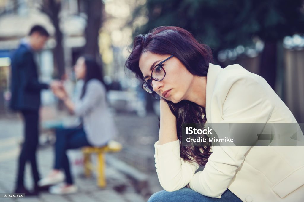 Young sad female sitting in the park Beautiful sad woman sitting alone and looking away from one happy couple on the next bench. Focus on the sad woman. Envy Stock Photo