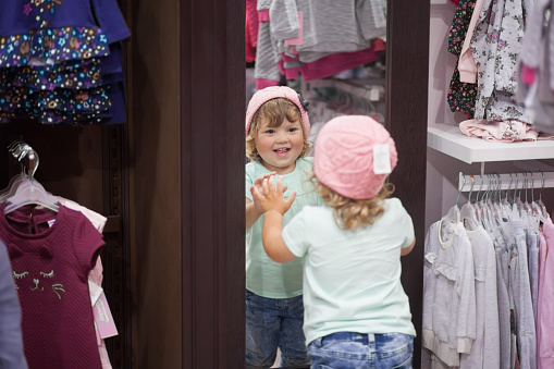 Adorable toddler girl in baby apparel store looking at the mirror, choosing clothes