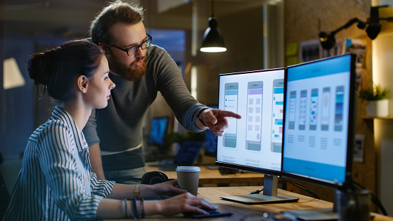 Female UX Architect Has Discussion with Male Design Engineer, They Work on Mobile Application Late at Night, She Drinks Coffee. In the Background Wall with Project Sticky Notes and Other Studio Employees.