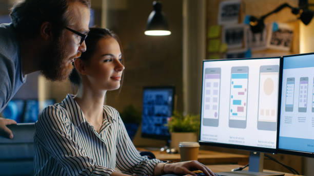 istock Female UX Architect Has Discussion with Male Design Engineer, They Work on Mobile Application Late at Night, She Drinks Coffee. In the Background Wall with Project Sticky Notes. 846843118