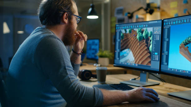 彼は 2 つの表示パソコンに取り組んで彼の木製のテーブルに座っている、創造的な男性のゲーム開発者が新しいレベル デザインのグラフィックと動作します。他の人々 はこの創造的なスタ� - computer programmer graphic designer graphic design studio designer ストックフォトと画像
