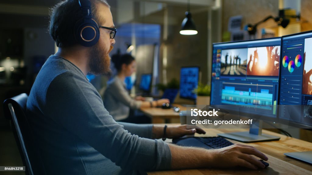 Male Videographer Edits and Cuts Footage and Sound on His Personal Computer, Puts on His Monitors/ Headphones. His Office is Modern and Creative Loft Studio. The Media Stock Photo