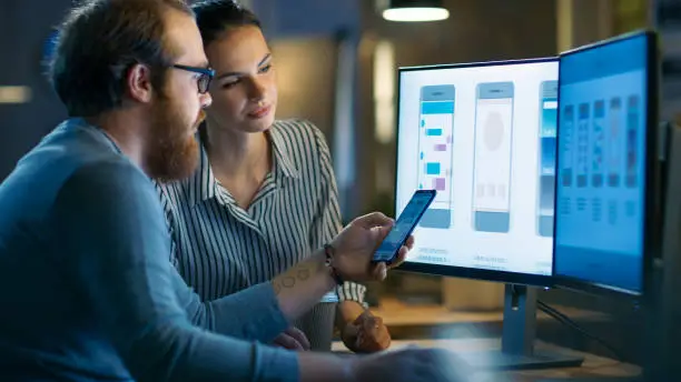 Photo of Handsome Male and Beautiful Female Mobile Application Designers Test and Discuss New App Features. They Work on a Personal Computer with Two Displays, in a Creative Office Space they Share with Other Talented People.