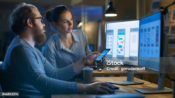 Handsome Male And Beautiful Female Mobile Application Designers Test And Discuss New App Features They Work On A Personal Computer With Two Displays In A Creative Office Space They Share With Other Talented People Stock Photo - Download Image Now