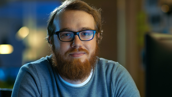 Joven había barbudo a hombre creativo con gafas piensa en un problema mientras trabaja en su computadora Personal. Mira sonriendo a la cámara. photo