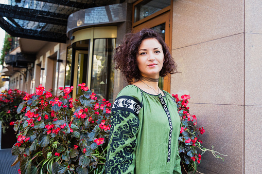 Lovely girl with short dark curly hair dressed in ethnic green embroidery dress standing near building. Accessories on the neck of the girl make the look modern and stylish. Embroidery, ethnic style, modern