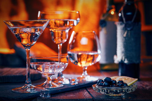 Liquids in various cocktail glasses in line. Olives and wine bottles on the table. Fire background