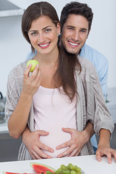 Cheerful man holding the belly of his pregnant wife Cheerful man holding the belly of his pregnant wife who is holding a green apple silver platter stock pictures, royalty-free photos & images