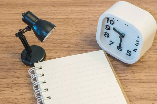 istock Closeup a note book with small lamp and white alarm clock on wood desk textured background 847104882