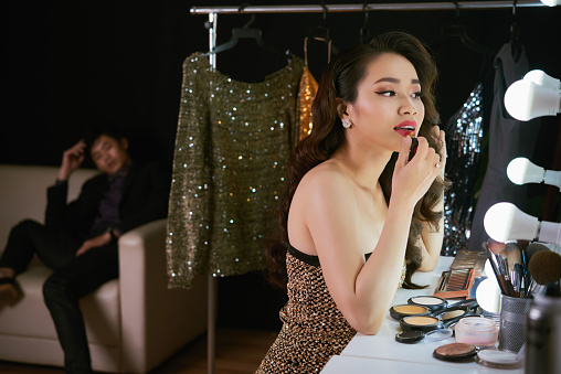 Gorgeous Vietnamese woman doing make-up, her tired boyfriend is waiting in the background