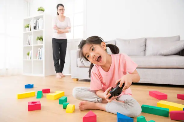 excited little girl playing joystick video game with colorful blocks toys scattered in living room wood floor and unhappy mom standing at rear angrily looking at her.