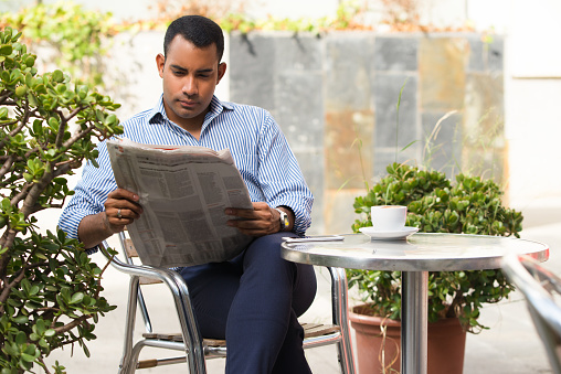 Serious Hispanic young man sitting in outdoor cafe, reading newspaper and drinking coffee. Confident executive manager reading recent news during lunchtime. Business news concept