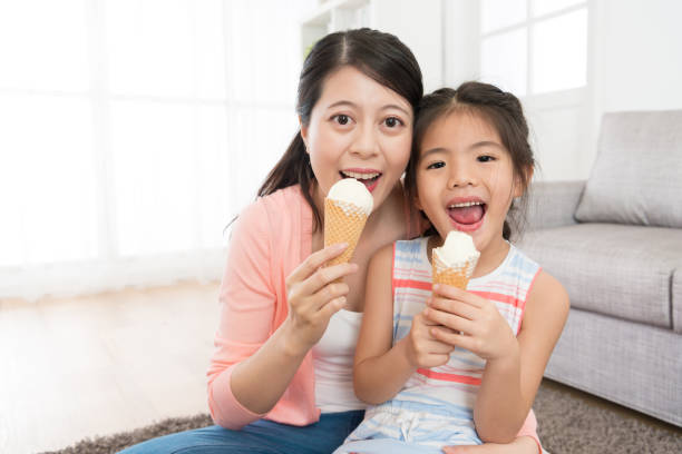 closeup photo of happy woman with little girl closeup photo of happy woman with little girl sitting on living room floor eating ice cream together and looking at camera laughing. asian little chinese girls eating ice cream stock pictures, royalty-free photos & images