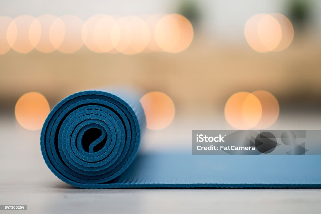 Calming Yoga A blue yoga mat is being rolled out onto a white floor inside a yoga studio. It is the evening and there are blurry candle lights behind the mat. Yoga Stock Photo